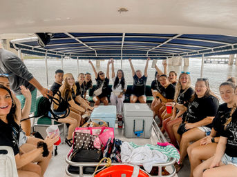 Cheerful group of friends in matching black shirts on a covered pontoon boat near a bridge, surrounded by coolers, bags, and drinks, raising hands and cups on a lively river boat party