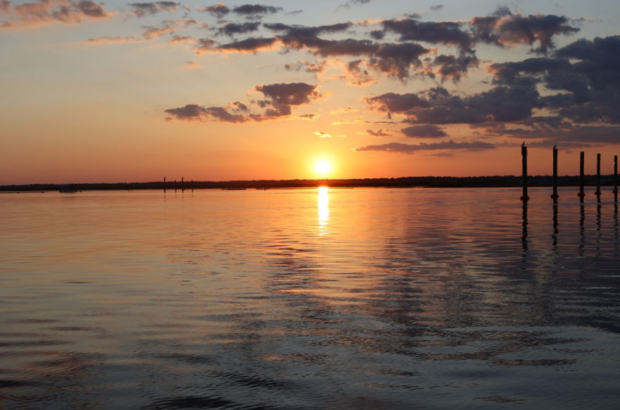 Tranquil golden sunset over a calm bay, sun reflecting on rippled water beneath scattered clouds with silhouetted pier pilings on the right