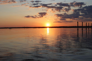 Tranquil golden sunset over a calm bay, sun reflecting on rippled water beneath scattered clouds with silhouetted pier pilings on the right