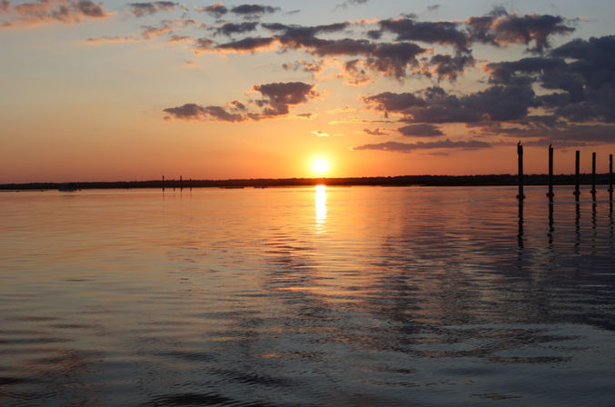 Tranquil golden sunset over a calm bay, sun reflecting on rippled water beneath scattered clouds with silhouetted pier pilings on the right