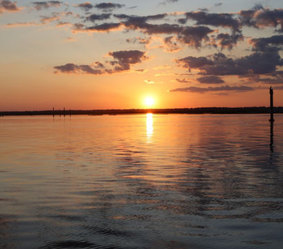 Tranquil golden sunset over a calm bay, sun reflecting on rippled water beneath scattered clouds with silhouetted pier pilings on the right