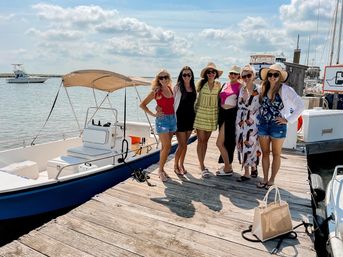 Six friends in summer outfits and sun hats posing on a wooden marina dock beside a small blue-and-white motorboat with a tan canopy, sunny sky and boats on the coastal harbor in the background, canvas tote on the dock.