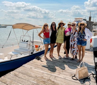Six friends in summer outfits and sun hats posing on a wooden marina dock beside a small blue-and-white motorboat with a tan canopy, sunny sky and boats on the coastal harbor in the background, canvas tote on the dock.