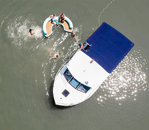 Aerial view of a white motorboat with blue canopy anchored in calm water, friends swimming and lounging on a crescent inflatable during a sunny summer boating day