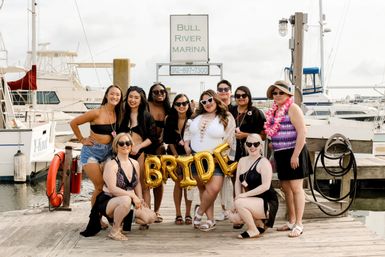 Bachelorette group of women on a marina dock posing with gold 'BRIDE' balloons, bride in a white swimsuit with heart sunglasses, surrounded by yachts and harbor pilings.