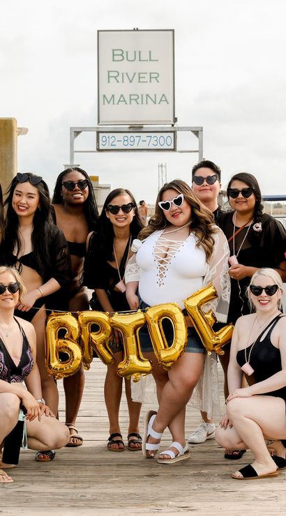 Bachelorette group of women on a marina dock posing with gold 'BRIDE' balloons, bride in a white swimsuit with heart sunglasses, surrounded by yachts and harbor pilings.