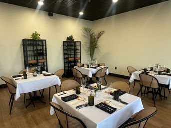 Cozy modern restaurant dining room with white-tablecloth tables set for dinner, rattan chairs, wine racks and a tall indoor palm under soft overhead lighting
