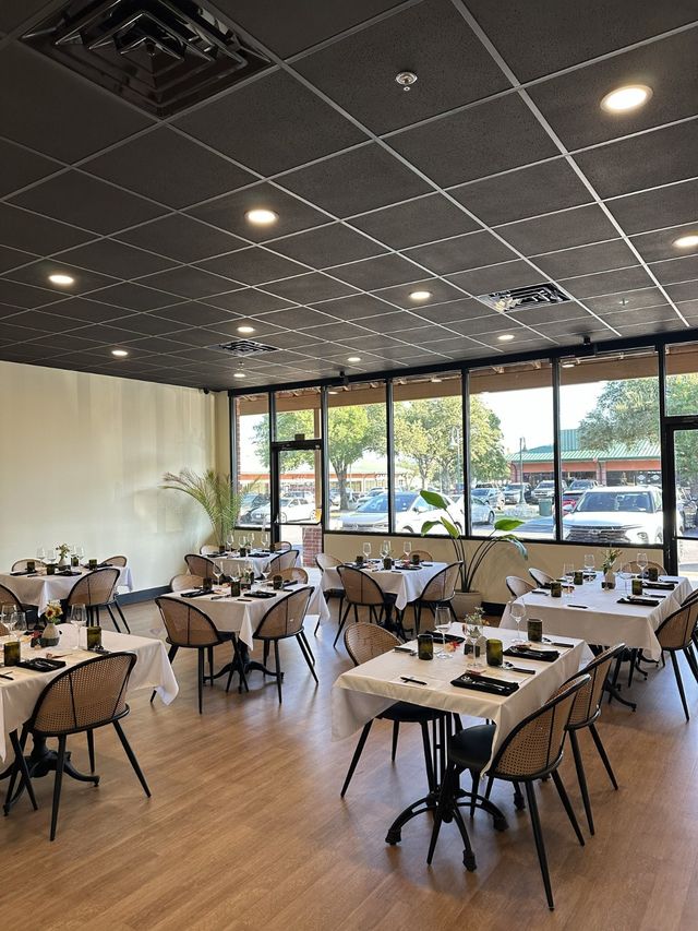 Bright suburban restaurant interior with white-tablecloth tables and rattan chairs set for dining, wood floors and large windows overlooking a parking lot and trees.