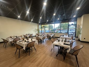 Bright modern restaurant dining room with wood floors and black ceiling, white-tablecloth tables set for guests, rattan chairs, potted plants and large street-side windows showing parked cars and trees.