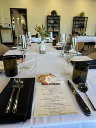 Autumn-themed restaurant table set for a wine tasting: white linen, wine glasses and recycled green tumblers, black napkins with silverware, printed menu and small floral vase, wine racks in the background.