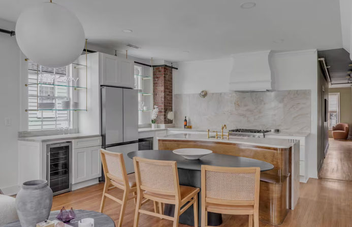 Bright modern open-plan kitchen with marble backsplash and white cabinetry, stainless steel refrigerator, brass faucet on a curved wooden island, black oval dining table with three cane-back chairs, exposed brick column, glass shelving and a large globe pendant light over hardwood floors.