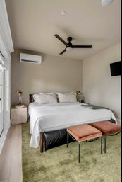 Cozy modern bedroom interior with king bed in white linens, ceiling fan, wall-mounted air conditioner, green area rug, two suede ottoman benches at the foot, bedside tables and a wall TV.