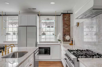 Bright contemporary kitchen with white marble countertops, brass faucet, stainless gas range, glass shelving and exposed brick accent.
