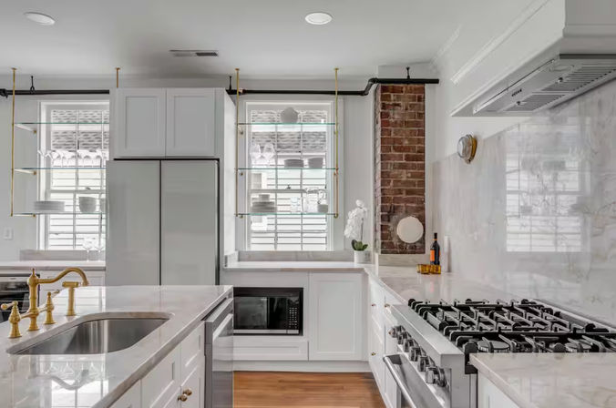 Bright contemporary kitchen with white marble countertops, brass faucet, stainless gas range, glass shelving and exposed brick accent.