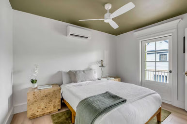 Sunlit modern bedroom with olive-green ceiling, white bedding and throw, wooden platform bed and burl-wood nightstands, orchid and lamp, wall-mounted AC, ceiling fan, and glass door to a small balcony overlooking neighboring houses.