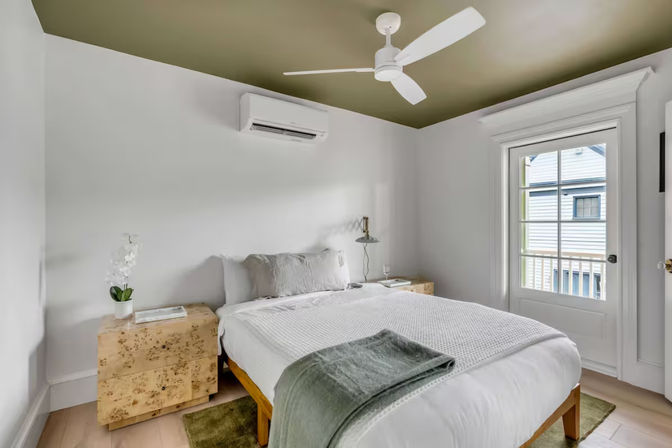 Sunlit modern bedroom with olive-green ceiling, white bedding and throw, wooden platform bed and burl-wood nightstands, orchid and lamp, wall-mounted AC, ceiling fan, and glass door to a small balcony overlooking neighboring houses.