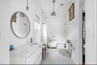 Airy all-white bathroom with freestanding clawfoot tub and shower, modern white vanity with round mirror, pendant lights, shuttered window, wooden bench and toilet.