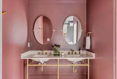 Chic mauve-pink bathroom interior with a double marble sink vanity, two oval mirrors, gold brass fixtures, towel ring and a vase of dark flowers