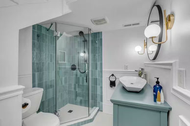 Modern bathroom with frameless glass corner shower lined in teal vertical subway tile, white toilet, teal vanity with white vessel sink, round mirror and brass globe wall sconces.