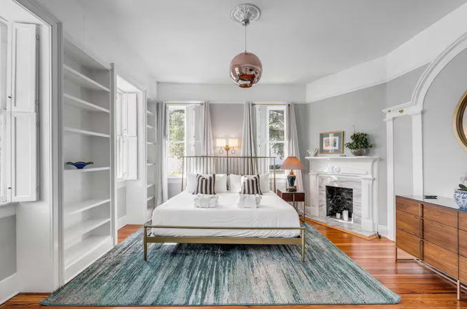 Sunlit historic bedroom with brass bed frame, teal area rug, hardwood floors, fireplace, built-in shelves and large windows