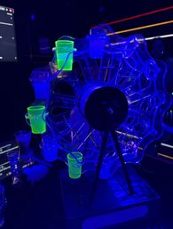 UV-lit rotating Ferris wheel shot glass carousel glowing neon green and blue on a bar counter under blacklight in a nightclub