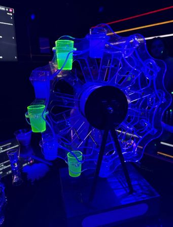 UV-lit rotating Ferris wheel shot glass carousel glowing neon green and blue on a bar counter under blacklight in a nightclub