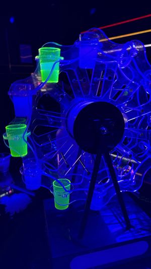 UV-lit rotating Ferris wheel shot glass carousel glowing neon green and blue on a bar counter under blacklight in a nightclub