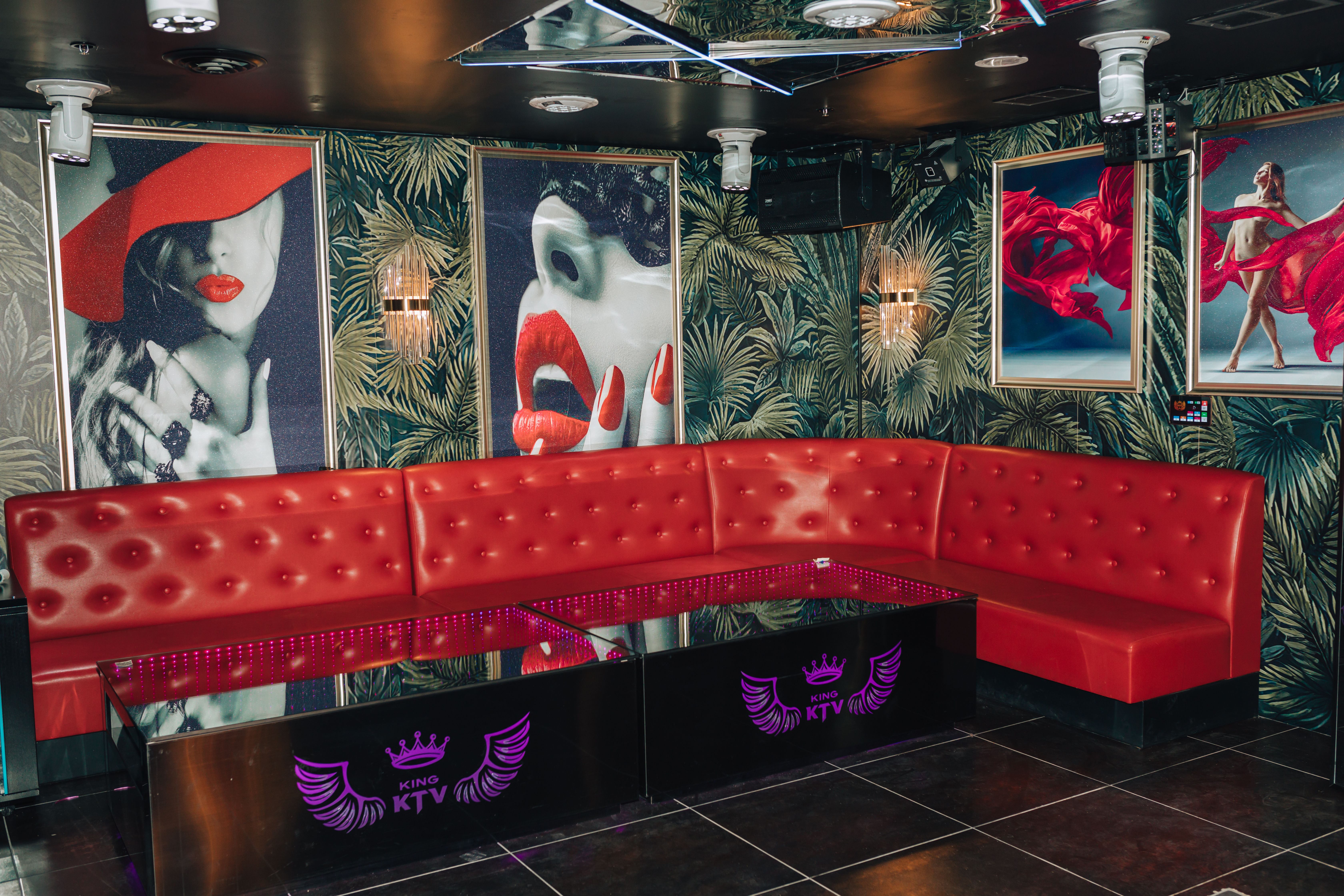 Vibrant KTV karaoke lounge interior with curved red tufted banquette, mirrored ceiling, tropical palm wallpaper, neon-lit glossy tables and pop-art posters featuring red lips and flowing fabric.