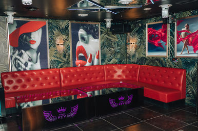 Vibrant KTV karaoke lounge interior with curved red tufted banquette, mirrored ceiling, tropical palm wallpaper, neon-lit glossy tables and pop-art posters featuring red lips and flowing fabric.