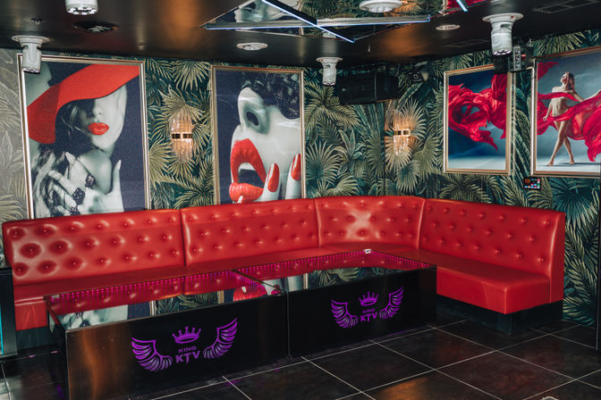 Vibrant KTV karaoke lounge interior with curved red tufted banquette, mirrored ceiling, tropical palm wallpaper, neon-lit glossy tables and pop-art posters featuring red lips and flowing fabric.