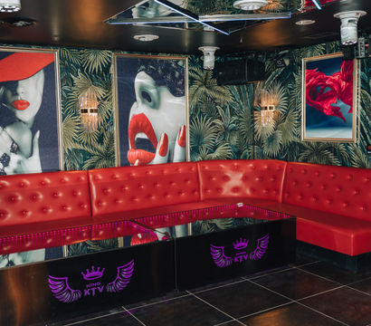 Vibrant KTV karaoke lounge interior with curved red tufted banquette, mirrored ceiling, tropical palm wallpaper, neon-lit glossy tables and pop-art posters featuring red lips and flowing fabric.