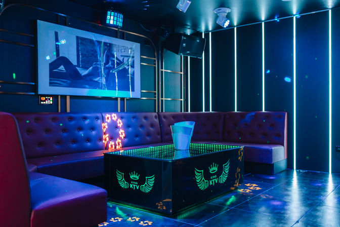 Modern neon-lit KTV karaoke room with tufted U-shaped booth, LED-lit glass table holding an ice bucket, wall screen and vertical light strips.