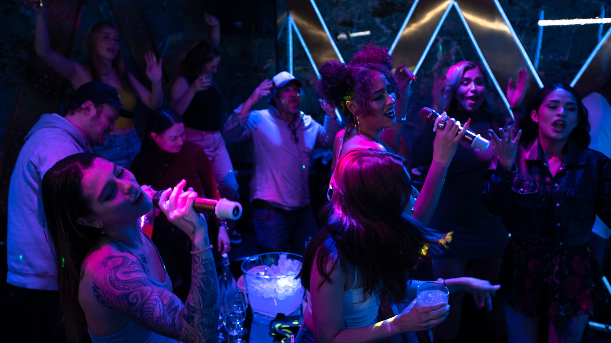 Lively karaoke night in a neon-lit bar — a crowd singing into microphones, holding drinks around a table with an ice bucket.