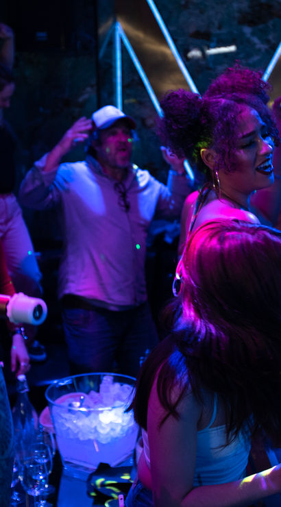 Lively karaoke night in a neon-lit bar — a crowd singing into microphones, holding drinks around a table with an ice bucket.
