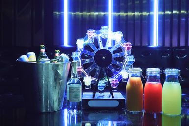 Neon-lit bar scene with a metal ice bucket of beer bottles and cans, a clear liquor bottle, an illuminated LED Ferris-wheel shot dispenser, and three glass bottles of colorful mixers on a reflective countertop.