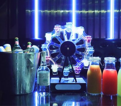 Neon-lit bar scene with a metal ice bucket of beer bottles and cans, a clear liquor bottle, an illuminated LED Ferris-wheel shot dispenser, and three glass bottles of colorful mixers on a reflective countertop.
