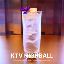 Tall whiskey highball cocktail in a clear glass with ice, citrus peel and purple edible flower garnish, sparkling soda in a warm-toned bar setting with blurred background
