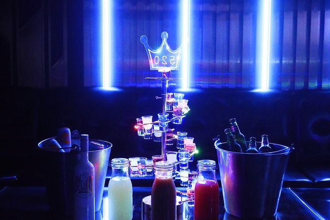 Blue neon-lit nightclub VIP table with glowing shot tower, bottle service in ice buckets and colorful mixers