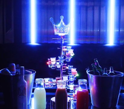 Blue neon-lit nightclub VIP table with glowing shot tower, bottle service in ice buckets and colorful mixers