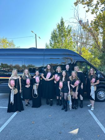 Playful group of about a dozen women in coordinated black outfits posing in front of a black party van in a sunny suburban parking lot, holding oversized cutout photos of a man's face with trees and blue sky behind them.
