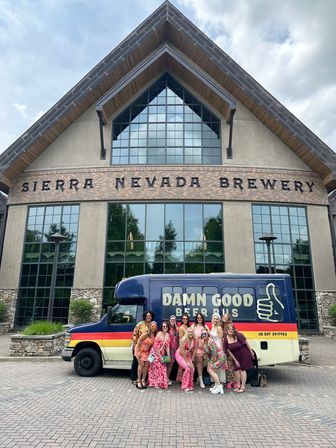Group of friends in bright outfits posing by a colorful beer tour bus outside a large A-frame craft brewery with brick, stone and tall mullioned windows.