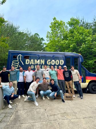Group of young men posing and smiling in front of a blue beer truck with a large thumbs-up logo and bold slogan, parked on a leafy driveway during an outdoor gathering.