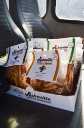 Cardboard tray of labeled artisanal pretzel stick bags from Asheville, NC with mustard and garlic and chive flavors, sitting on a vehicle seat.