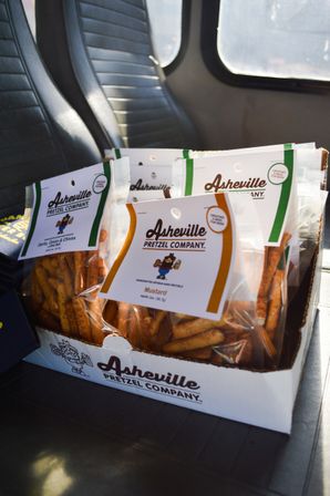 Cardboard tray of clear bags filled with flavored pretzel sticks (mustard and garlic) sitting on a van or bus seat by a window