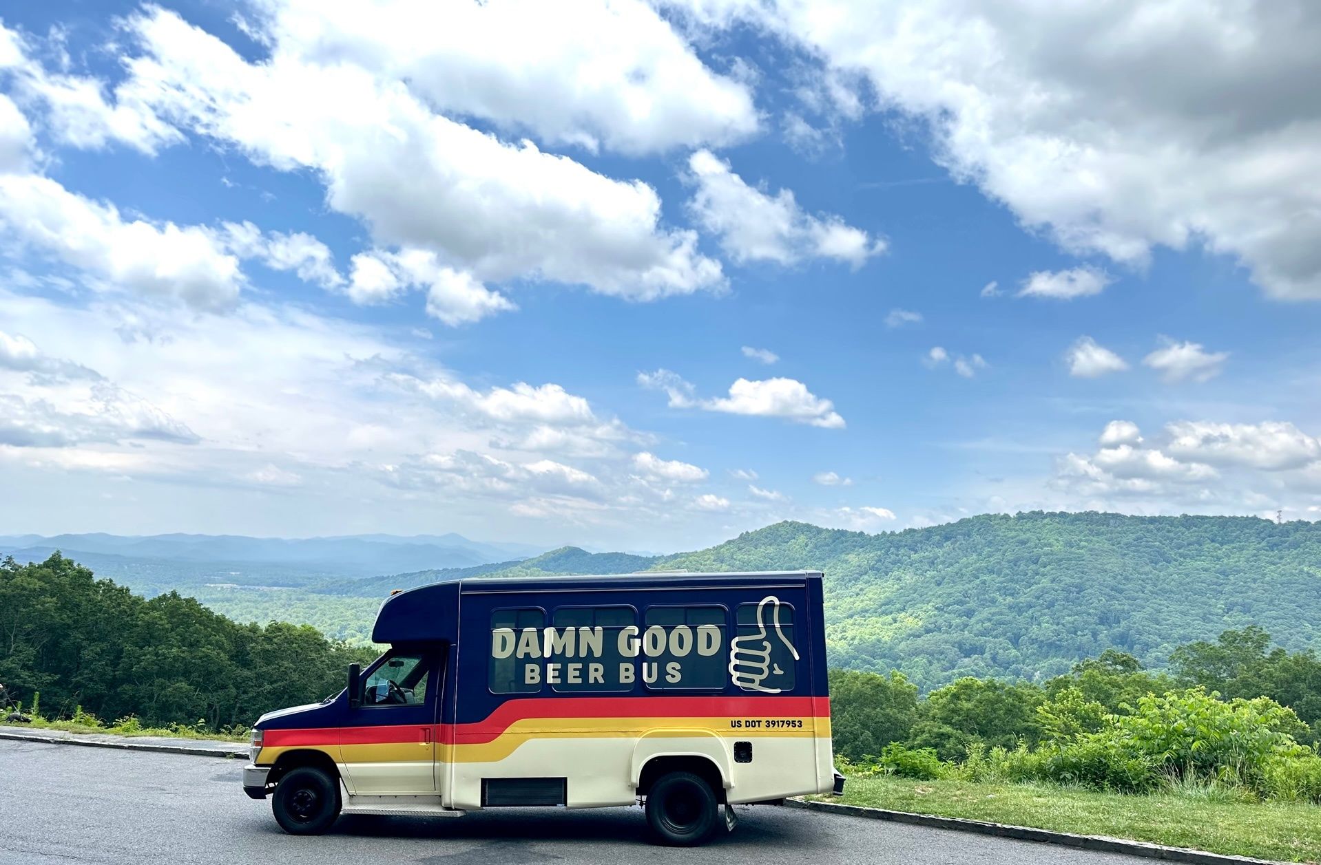 Colorful beer shuttle van with thumbs-up logo parked at a mountain overlook, overlooking rolling green hills under a bright blue sky dotted with fluffy clouds.