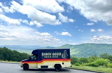 Retro striped beer bus parked at a scenic mountain overlook with rolling green hills and distant blue ridges beneath a bright blue sky dotted with fluffy clouds.
