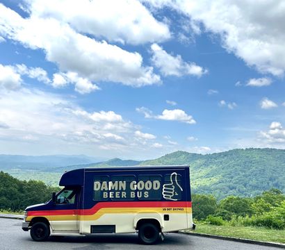 Colorful beer shuttle van with thumbs-up logo parked at a mountain overlook, overlooking rolling green hills under a bright blue sky dotted with fluffy clouds.