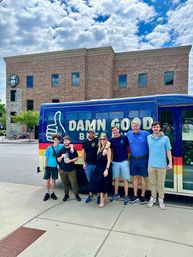 Seven people smiling and posing beside a colorful shuttle bus with a thumbs-up graphic in front of a brick office building under a bright blue sky.