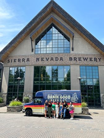 Group of friends posing by a colorful beer tour shuttle outside an alpine-style craft brewery with a large triangular roof, brick and stone facade and tall gridded windows on a sunny day