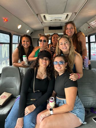 Seven smiling friends enjoying a party-bus ride, posing with drinks inside a shuttle with gray vinyl seats and city windows visible.